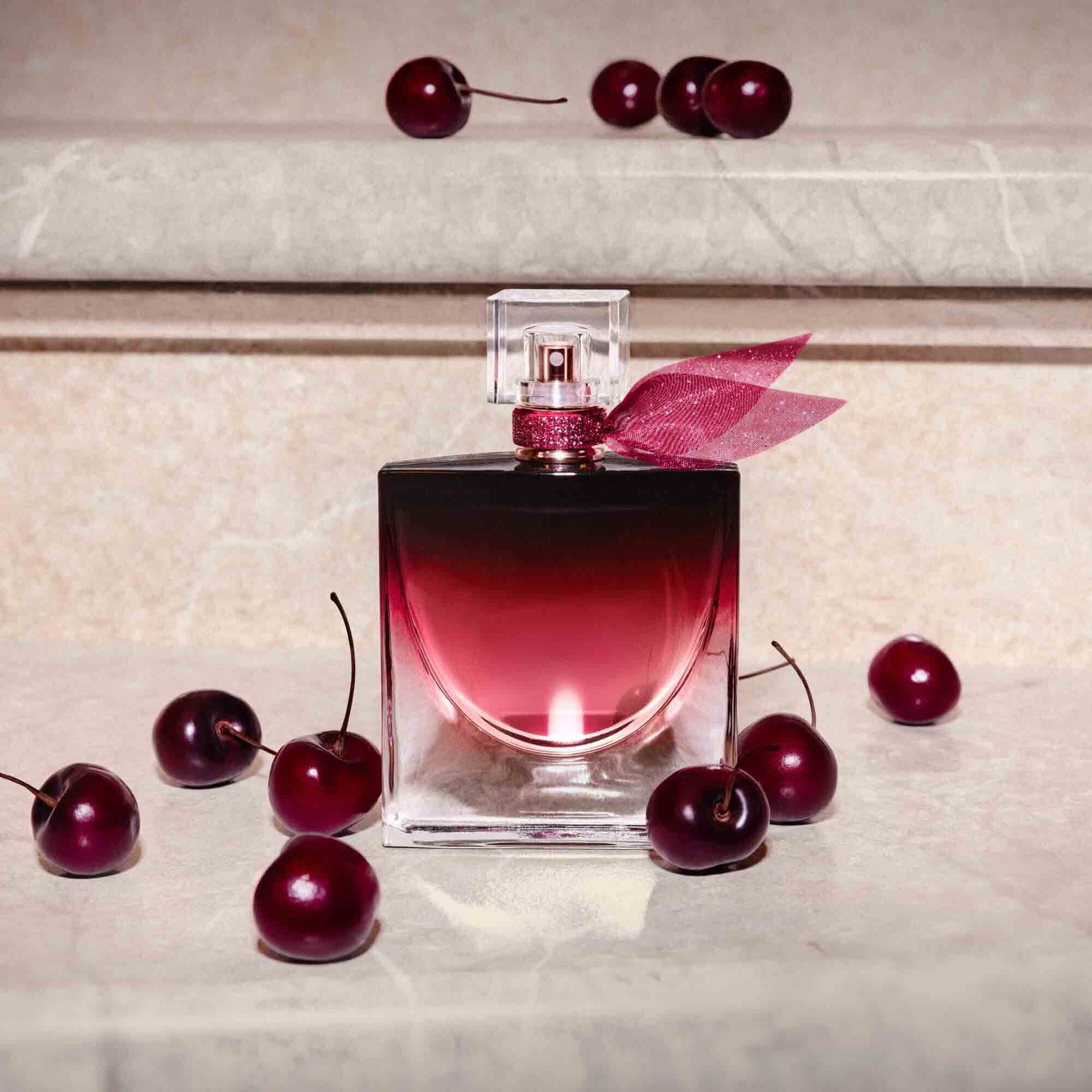 The burgundy perfume bottle stands on a stone ledge surrounded by several ripe, dark cherries.	