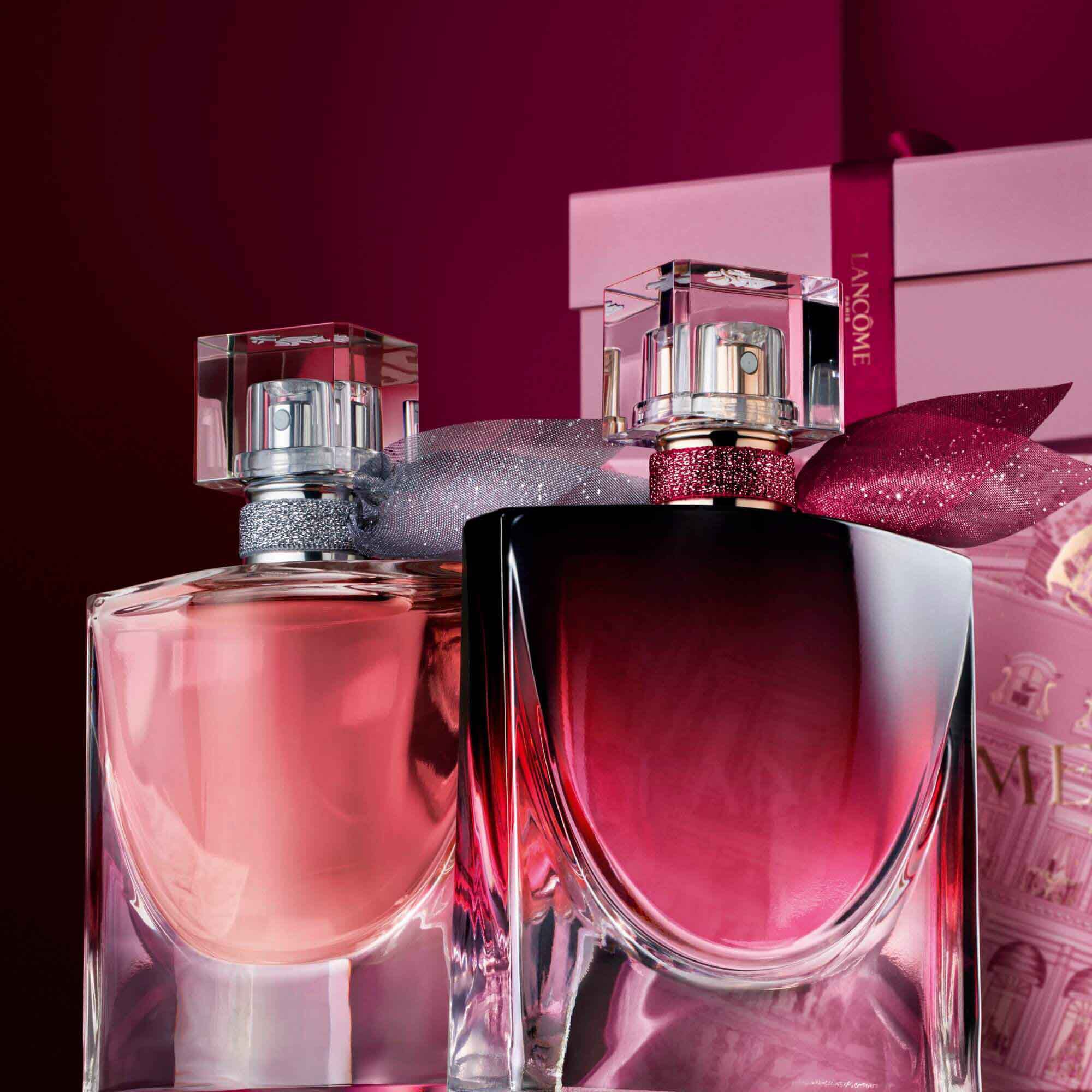 Image of La vie est belle EDP and La vie est belle L'Elixir Very Cherry standing in front of a pile of pink and dark red gift boxes, set against a dark red background. 