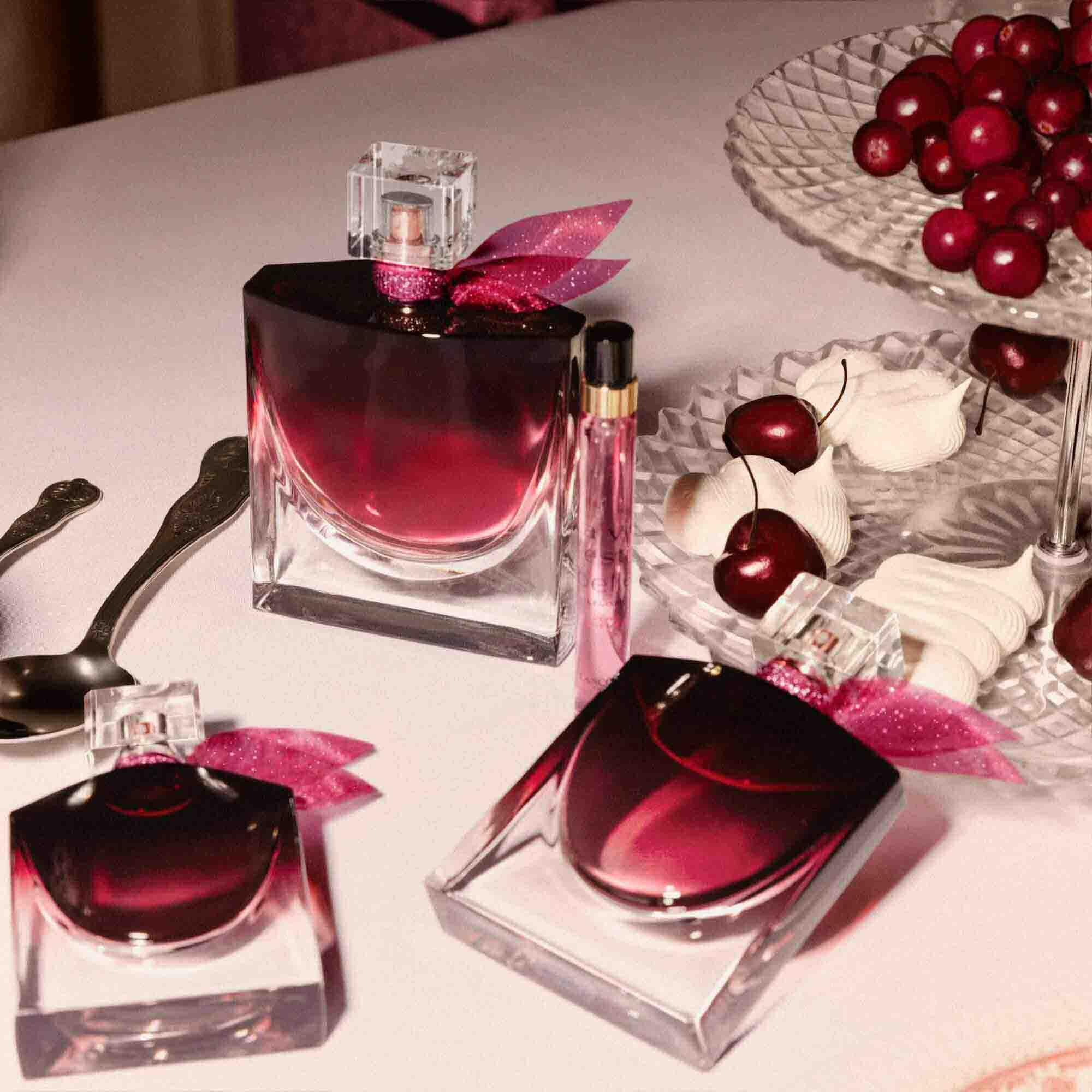 An elegant tabletop scene featuring multiple sizes of the perfume bottle alongside a bowl of fresh cherries and silver spoons.	